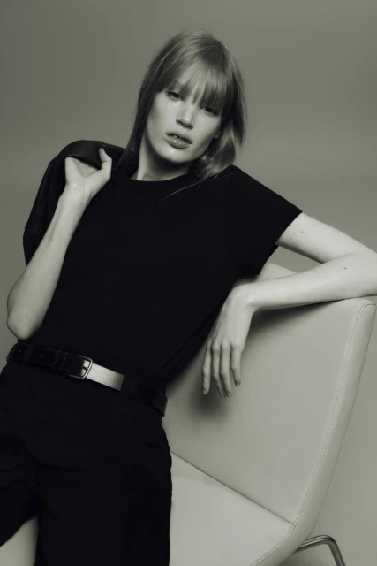 Blonde woman in black t-shirt and pants poses in studio against grey backdrop with jacket slung over shoulder while sitting in chair.