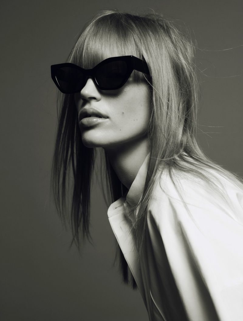 Monochrome close-up shot of a young adult woman wearing black sunglasses and a white blouse, looking to the side against a grey backdrop.