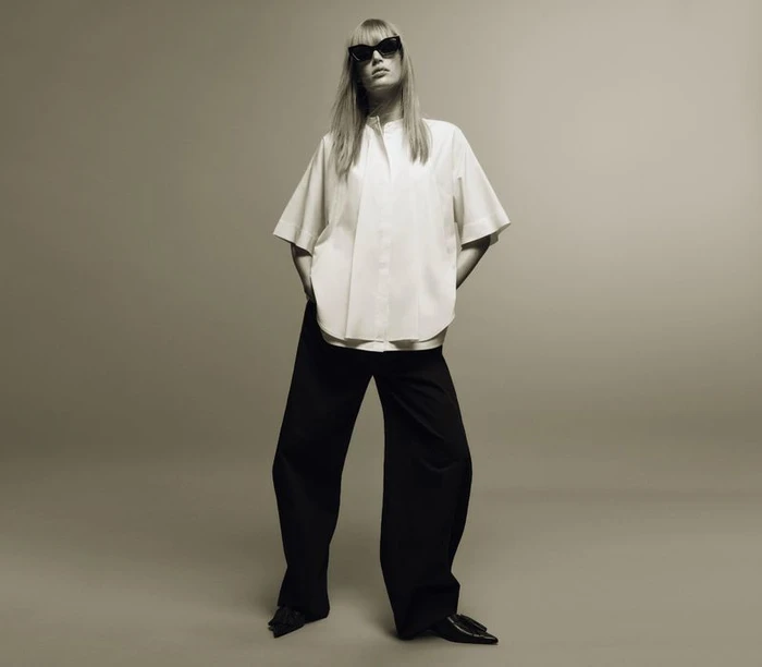 Model standing against a plain grey background wearing a white short-sleeve shirt, black trousers, and dark sunglasses.