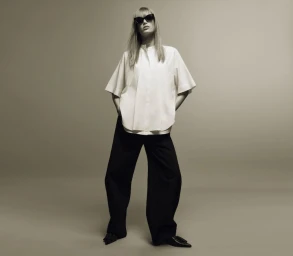 Model standing against a plain grey background wearing a white short-sleeve shirt, black trousers, and dark sunglasses.