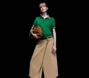 Model in green polo shirt and tan skirt holding a brown suede bag against a black background for COS Spring/Summer 25