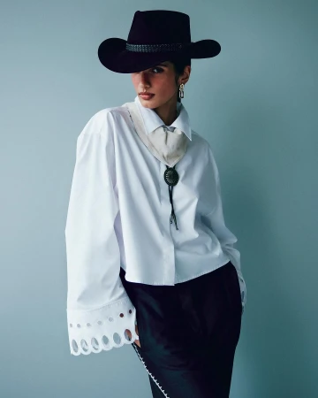 Fashionable woman with confident expression wears a cowboy hat, white collared shirt, bolo tie, and dark skirt, posing against a blue-gray backdrop.