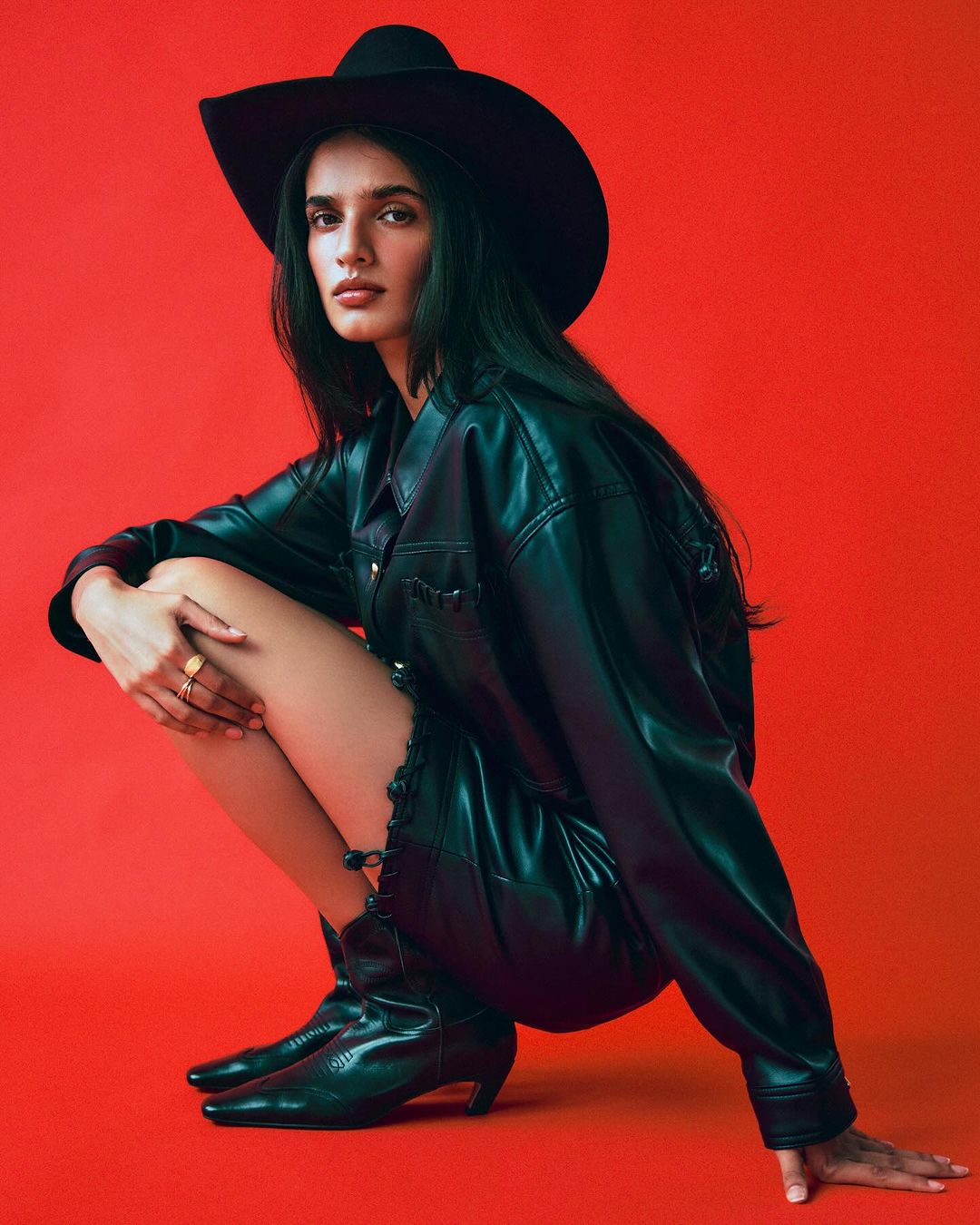 A model in a black leather jacket, hat, and cowboy boots poses against a vibrant red backdrop in a fashion-forward studio shot.