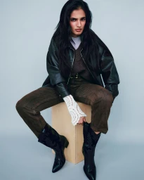 Anjali Torvi seated on a wooden box wearing a dark leather jacket, brown denim jeans, and black cowboy boots against a seamless grey backdrop.