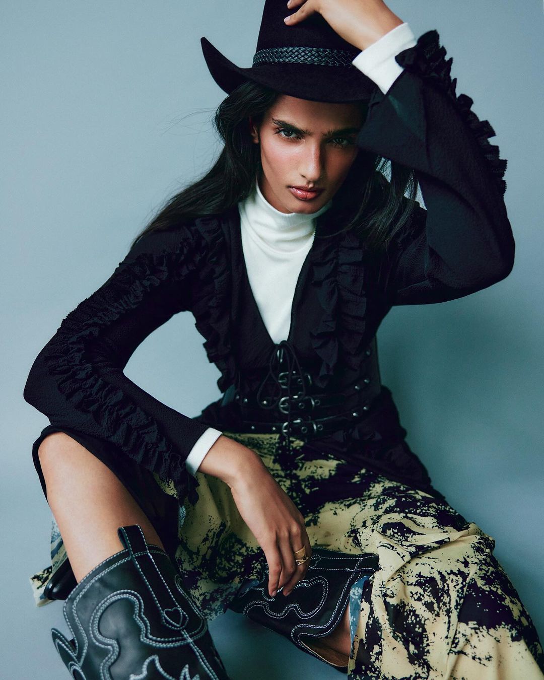 Woman in black cowboy hat and jacket poses against a gray background wearing patterned skirt and cowboy boots in a fashion editorial shot.
