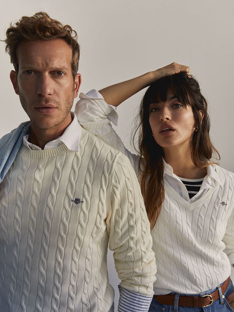 A fashionable couple poses indoors wearing cable-knit sweaters. The man stands confidently; the woman rests her hand on her head.