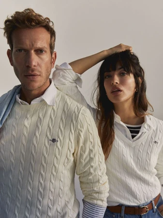 A fashionable couple poses indoors wearing cable-knit sweaters. The man stands confidently; the woman rests her hand on her head.