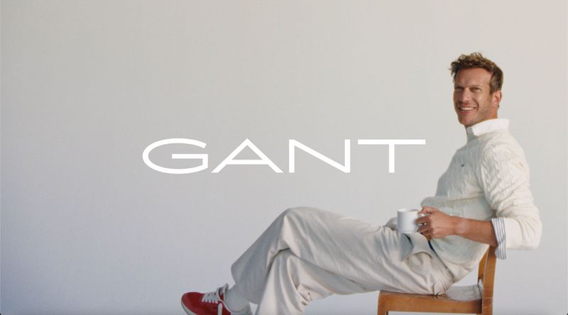 Man in a cable-knit sweater sits in front of a white background, holding a mug, with the GANT logo visible in the background.