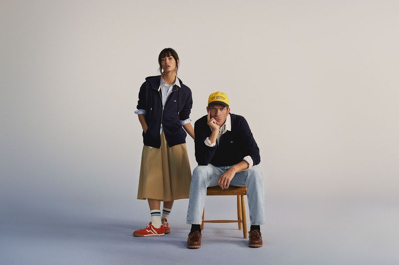 A stylish couple poses in a studio. The man sits on a wooden chair, wearing jeans and a navy sweater. The woman stands in a skirt and jacket.