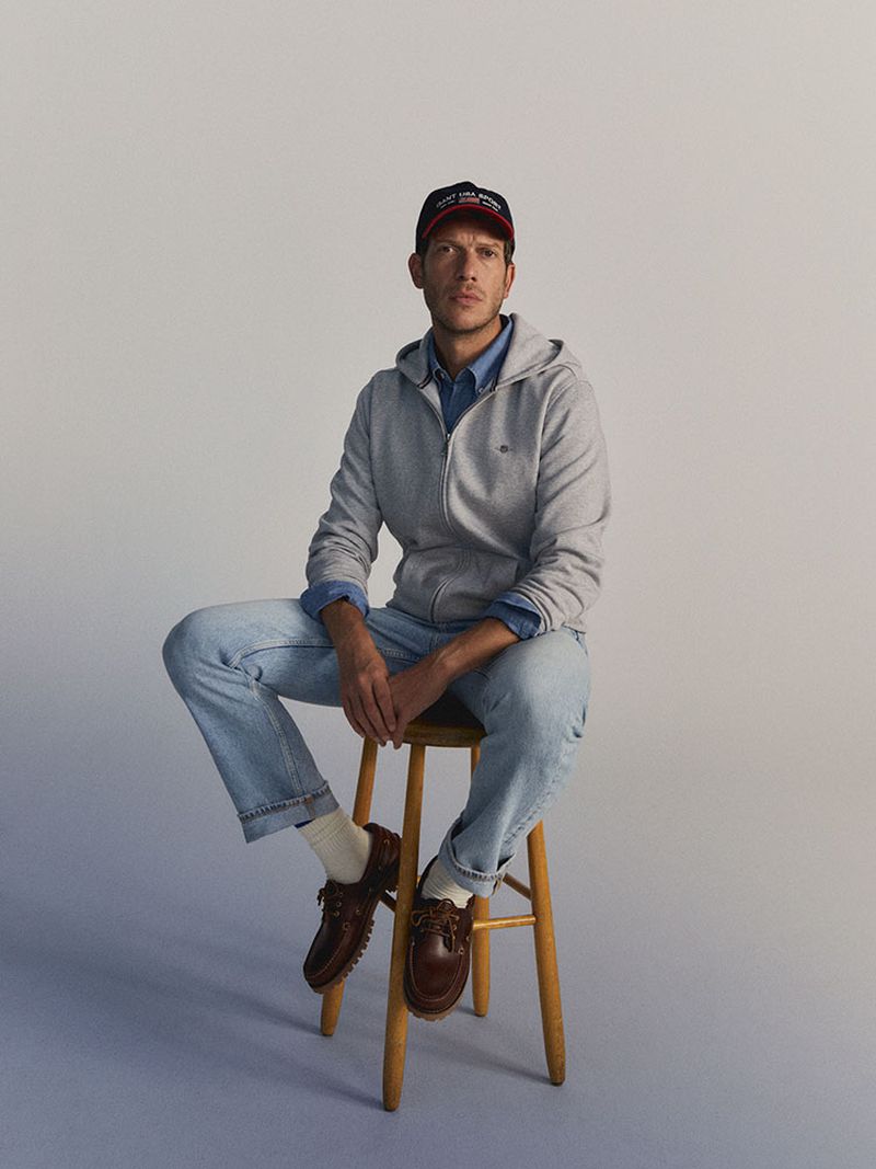 A man sitting on a wooden stool with a gray zip-up hoodie, blue jeans, brown shoes, and a baseball cap in a bright studio setting.