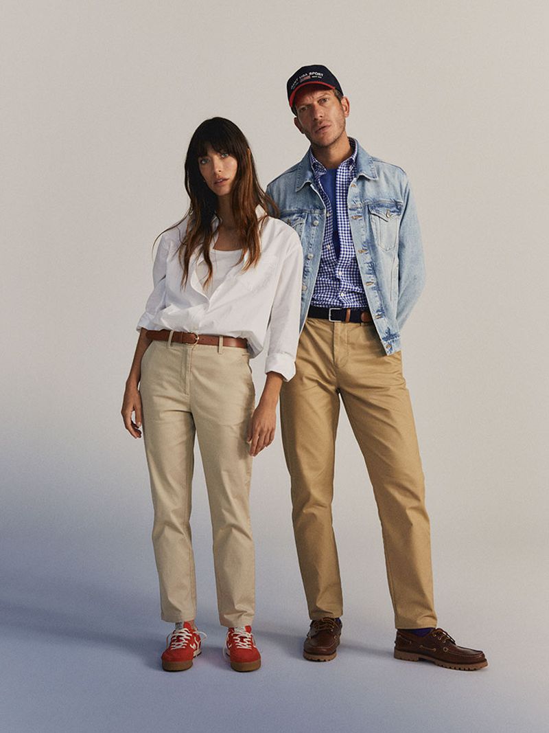 A casually dressed couple poses in a studio. The woman wears a white button-down, and the man wears a denim jacket and baseball cap.