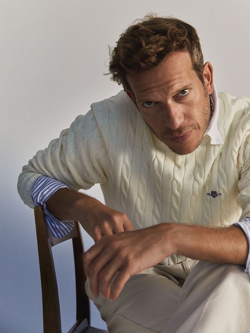 Portrait of a man wearing a cable-knit sweater and light-colored pants, seated on a wooden chair in front of a neutral background.