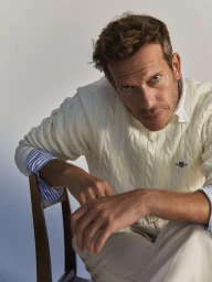 Man wearing a cream cable-knit sweater over a striped button-down shirt, seated on a wooden chair against a plain light background