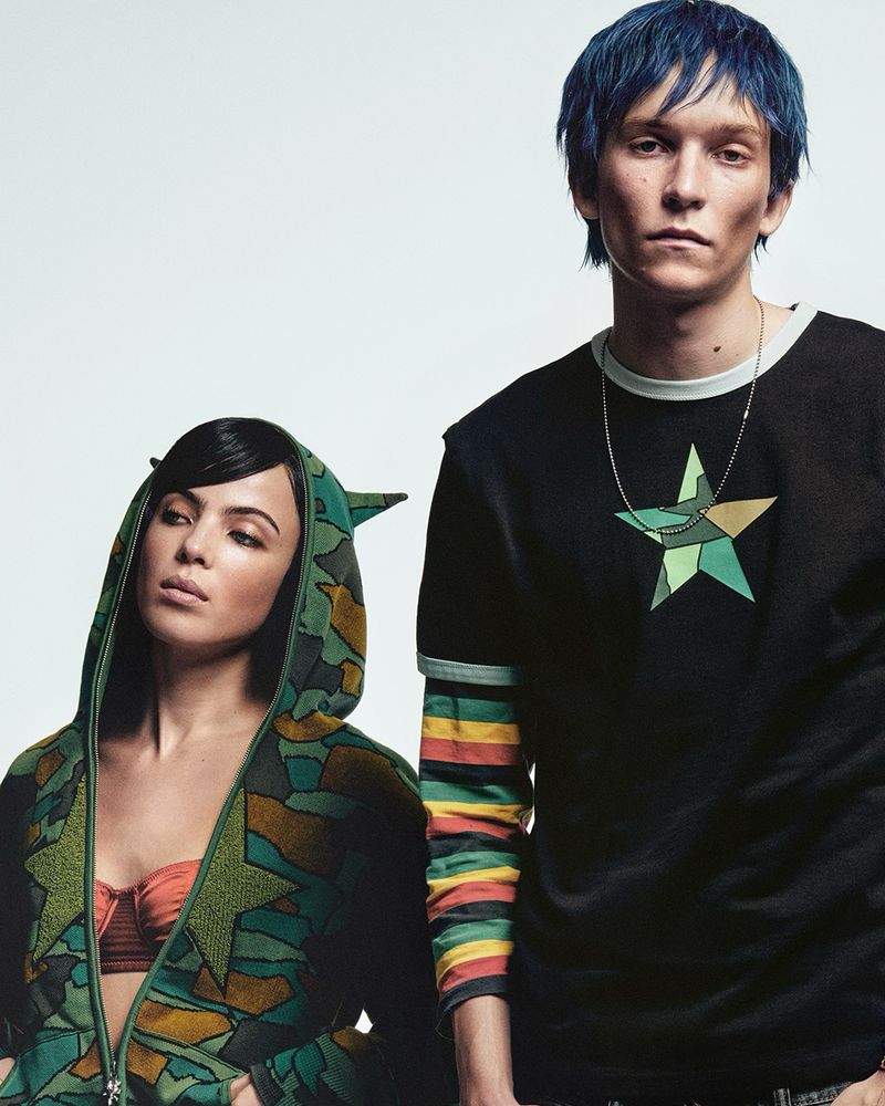 Two models pose in front of a white background, one with blue hair wearing a star-themed shirt, the other in a patterned hooded jacket.