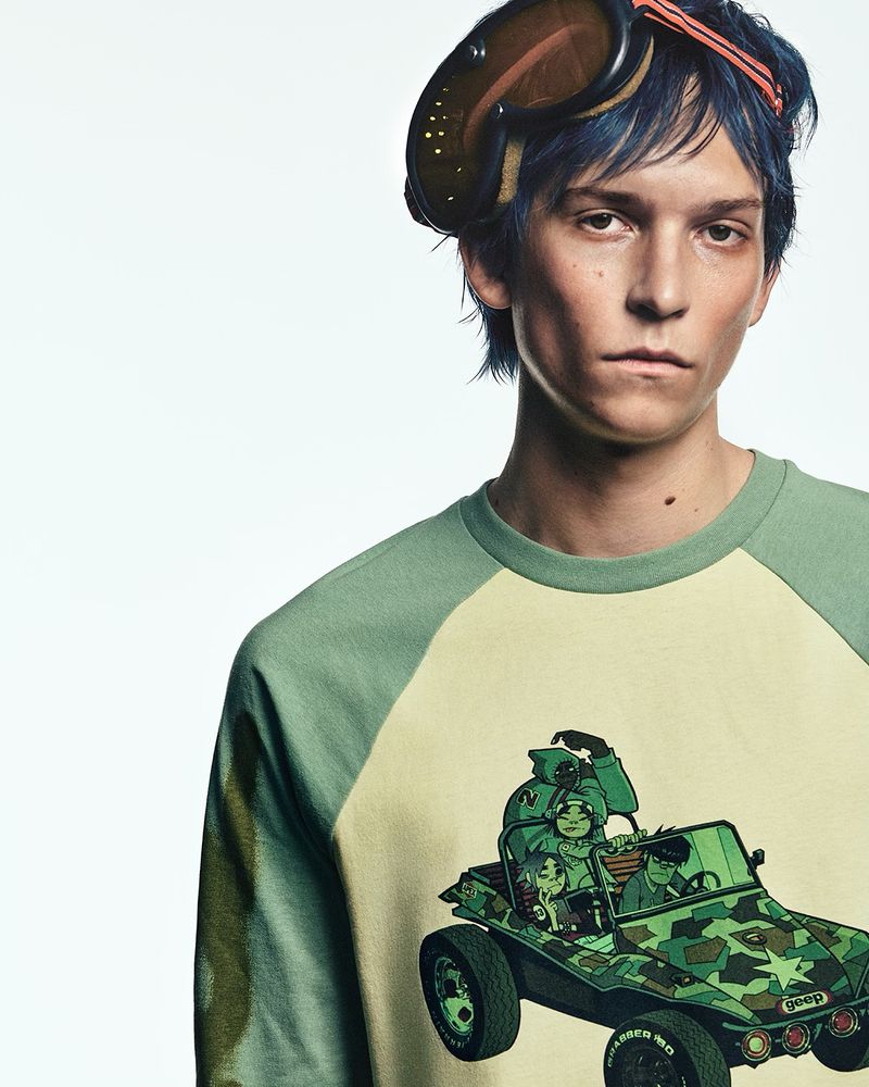 Adult male poses indoors wearing a printed shirt and goggles; a graphic of characters in a vehicle is printed on the garment.