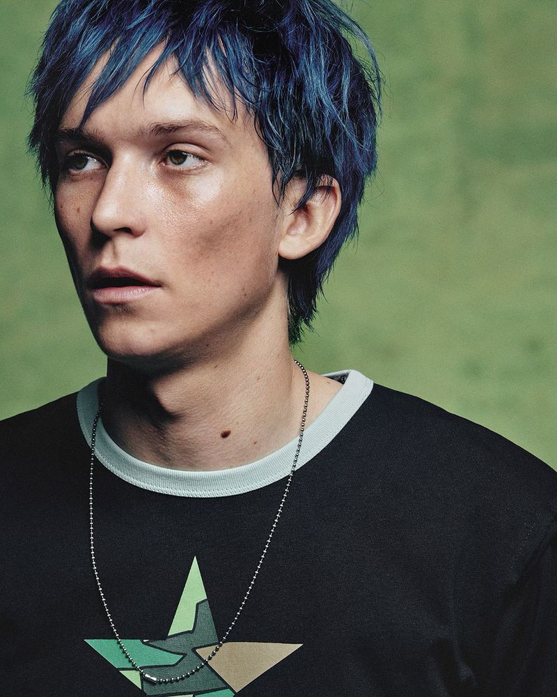 Portrait of a young adult male with blue hair wearing a black tee shirt with a star graphic against a light green background.