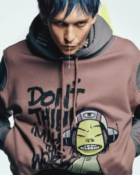 Model with blue hair wearing a brown graphic hoodie featuring Gorillaz artwork against a white studio background.