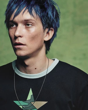 Close-up portrait of a male model with vibrant blue hair wearing a black star-print t-shirt against a textured green background.