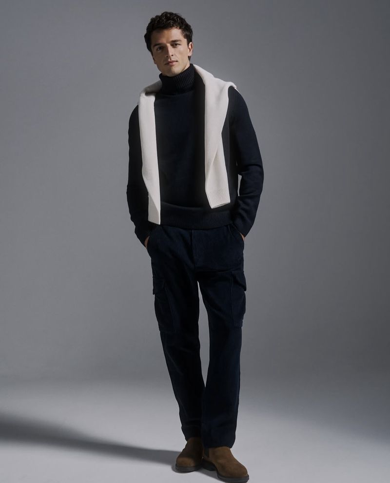 Full-length shot of a handsome adult male model in a black turtleneck sweater and black pants, with a white sweater draped over his shoulders.