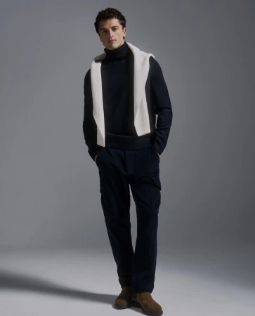 Full-length shot of a handsome adult male model in a black turtleneck sweater and black pants, with a white sweater draped over his shoulders.