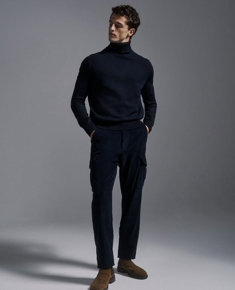 Full-length shot of a male model in a navy turtleneck, cargo pants, and brown boots standing in a photo studio with soft lighting.
