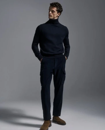Full-length shot of a male model in a navy turtleneck, cargo pants, and brown boots standing in a photo studio with soft lighting.