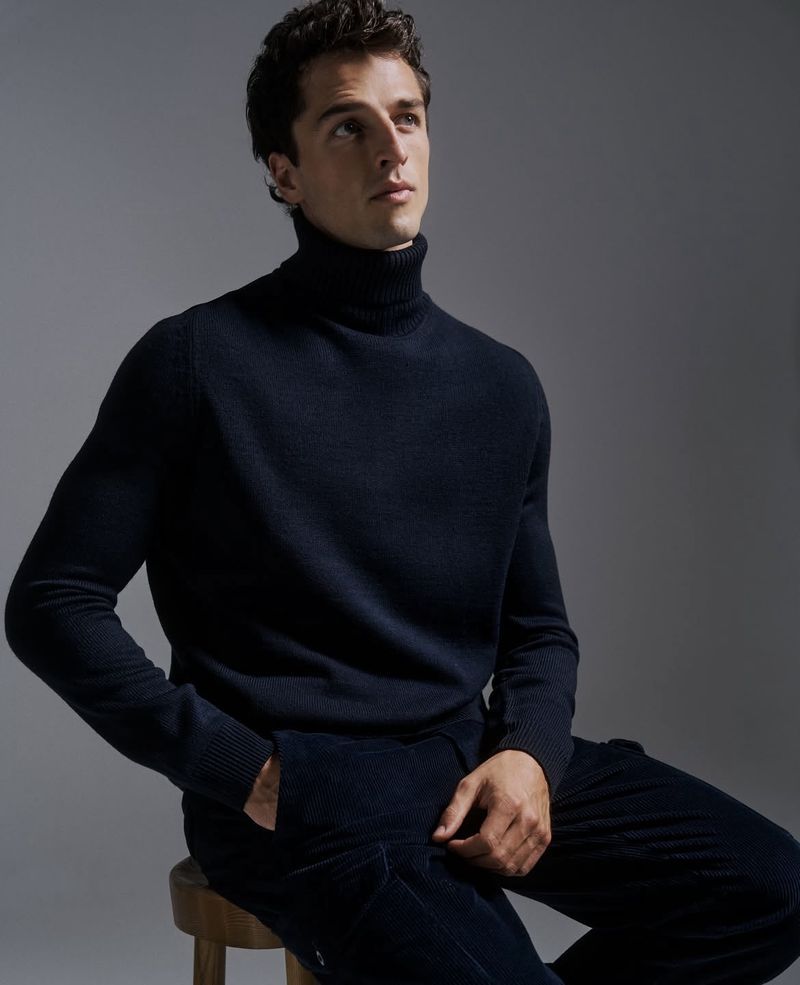 Portrait of a young adult male sitting on a stool in a navy blue turtleneck and pants, illuminated by soft light against a gray backdrop.
