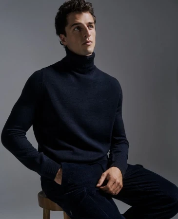 Portrait of a young adult male sitting on a stool in a navy blue turtleneck and pants, illuminated by soft light against a gray backdrop.