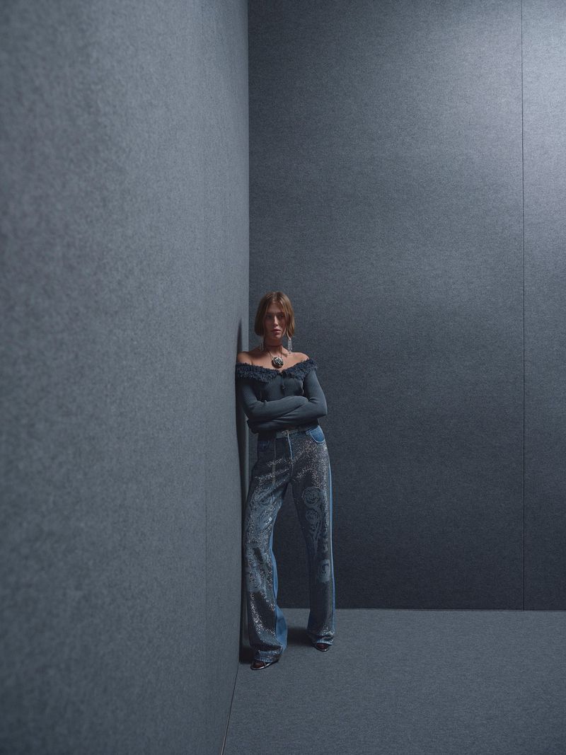 Full-length shot of a young woman with brown hair in a long-sleeved, off-the-shoulder top and sequined pants leaning against a gray wall.