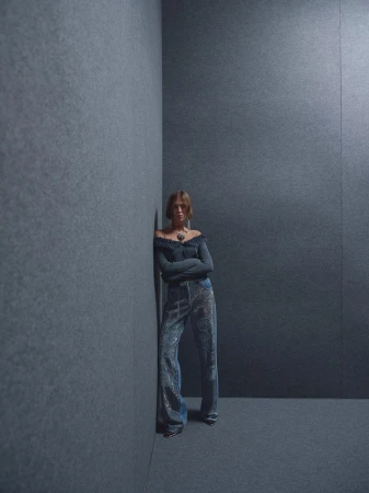 Full-length shot of a young woman with brown hair in a long-sleeved, off-the-shoulder top and sequined pants leaning against a gray wall.