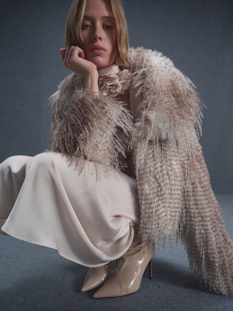 A fair-skinned young adult woman with blonde hair poses, wearing a feathered coat, cream dress, and high heels against a grey backdrop.