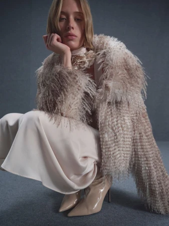 A fair-skinned young adult woman with blonde hair poses, wearing a feathered coat, cream dress, and high heels against a grey backdrop.