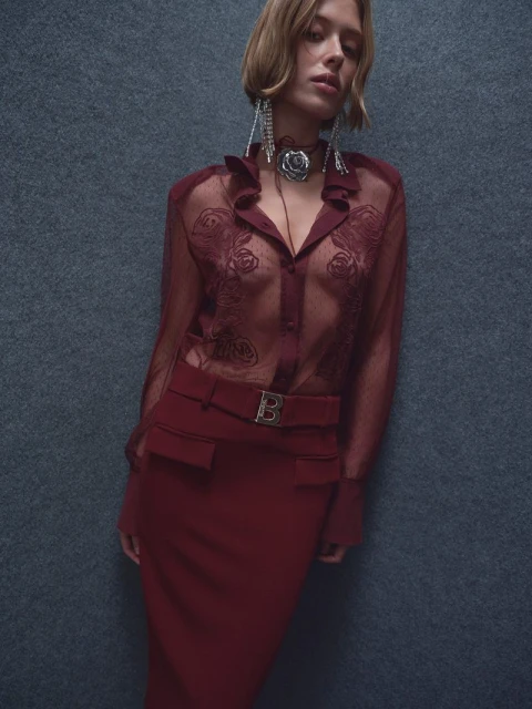 A fashionable woman poses in a wine-colored, sheer blouse with rose patterns and a matching skirt, complemented by statement jewelry.
