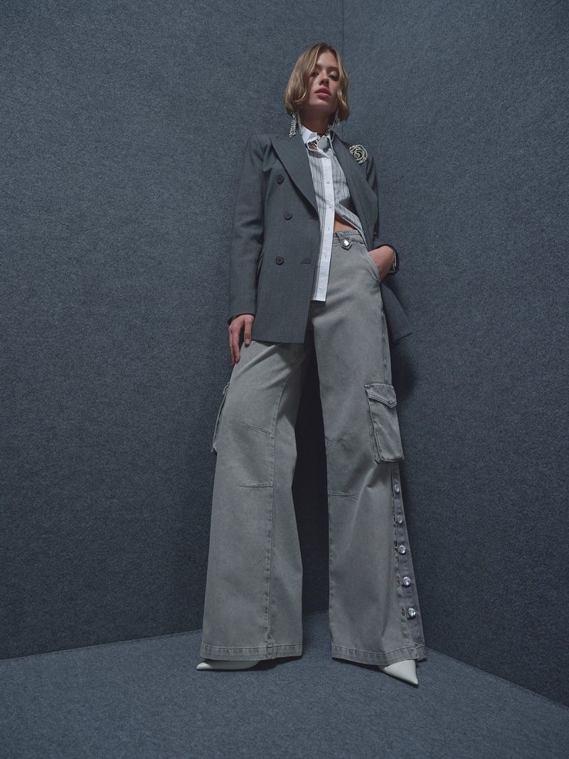 Woman in grey blazer, white blouse, wide-leg jeans, and white heels in a studio setting, fashion portrait, confident expression.