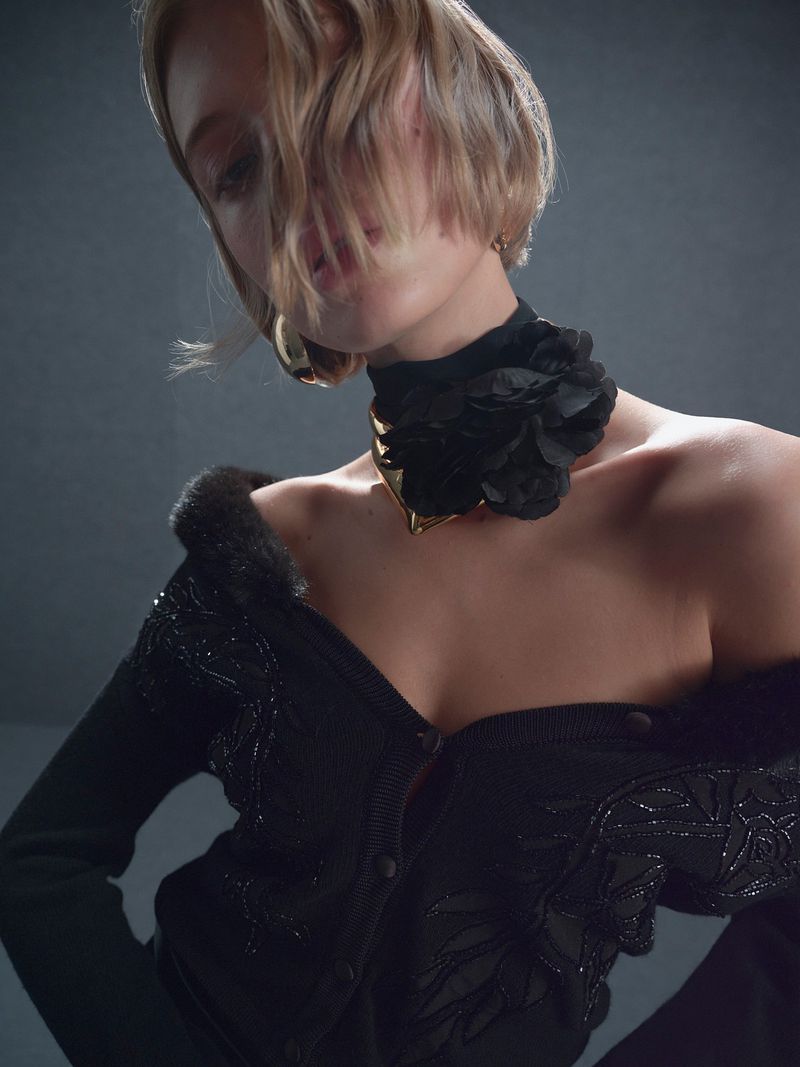 Fashion portrait of a woman wearing a black flower choker and beaded cardigan, posing in front of a neutral grey studio background.