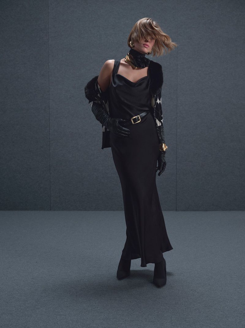 A stylish adult female fashion model in a black slip dress and jacket poses in front of a muted grey background in a studio setting.