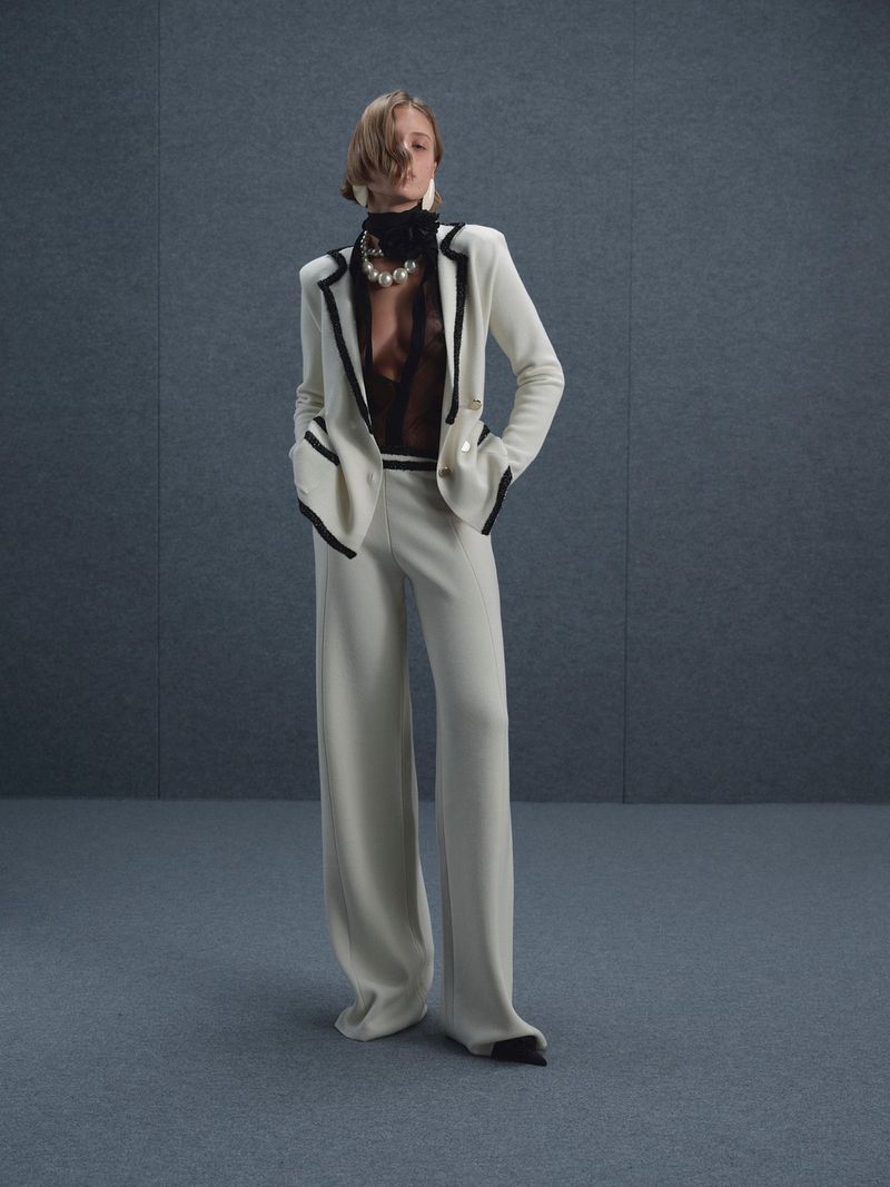 Model wearing a white suit jacket with dark trim and wide-leg trousers against a grey studio backdrop