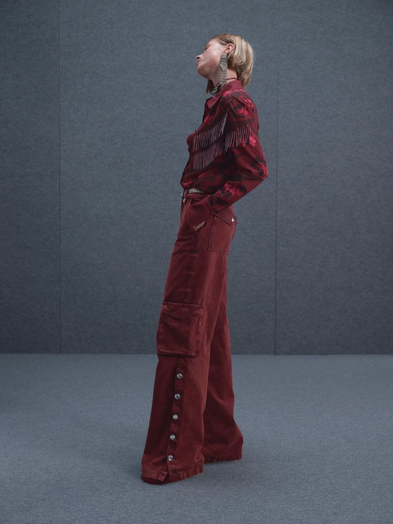 Full-length studio shot of a young adult female model wearing a button-down fringe shirt and wide-leg cargo pants in red-brown.