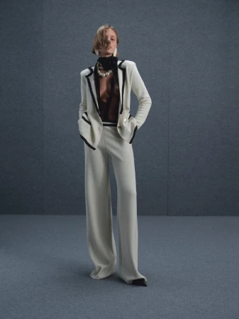 Model wearing a white suit jacket with dark trim and wide-leg trousers against a grey studio backdrop