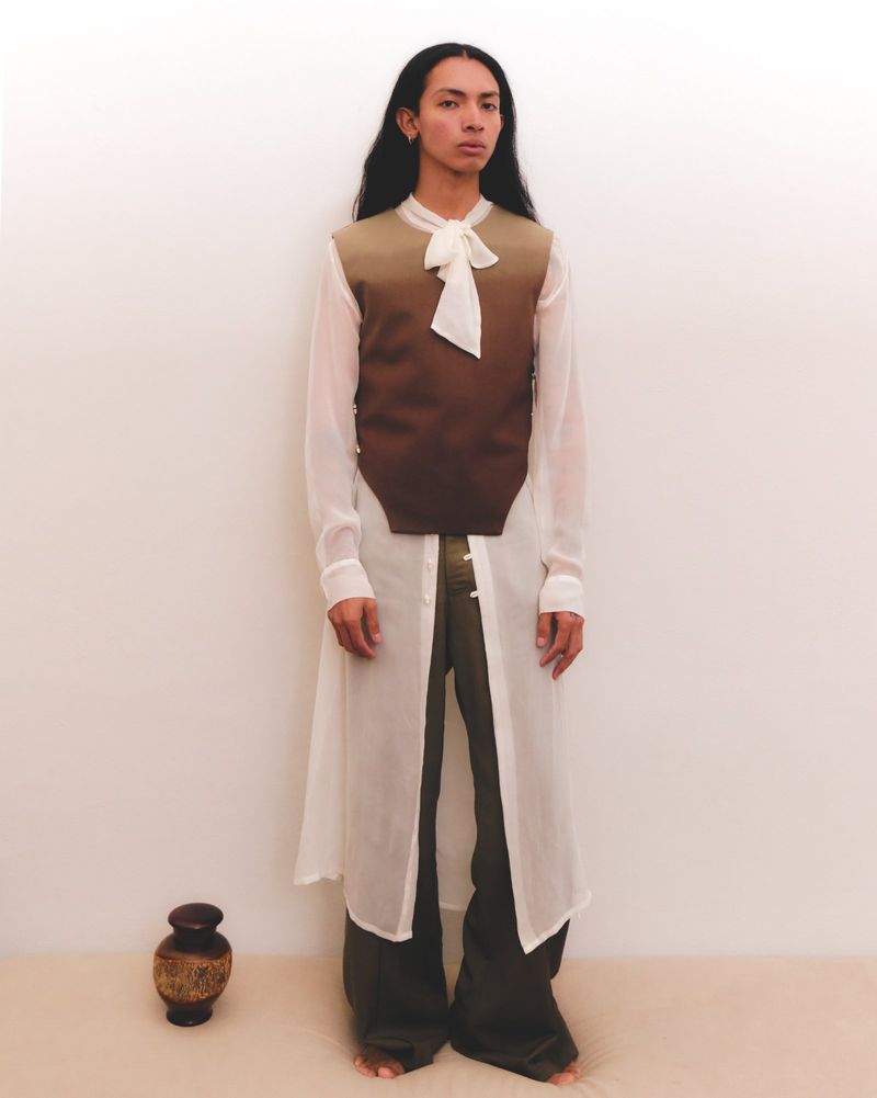 Full-length shot of a person with long hair, wearing a sheer white tunic, brown vest, and dark pants, standing barefoot indoors.