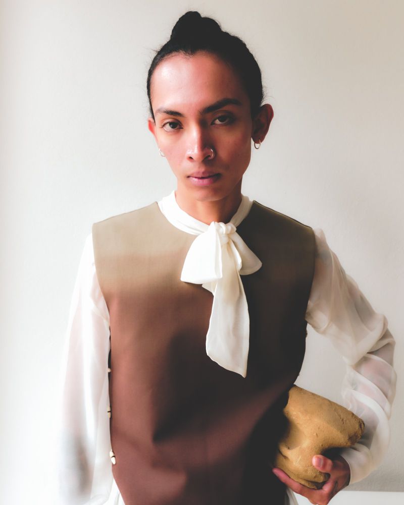 Portrait of person in ombré top with sheer sleeves and ivory bow tie holding beige object against neutral backdrop in studio light.