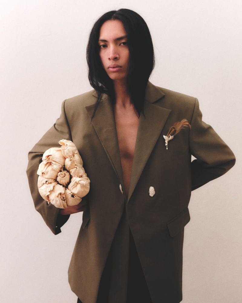 Elegant adult male model with long dark hair, wearing an olive green jacket, holding a garlic bunch against a white studio backdrop.