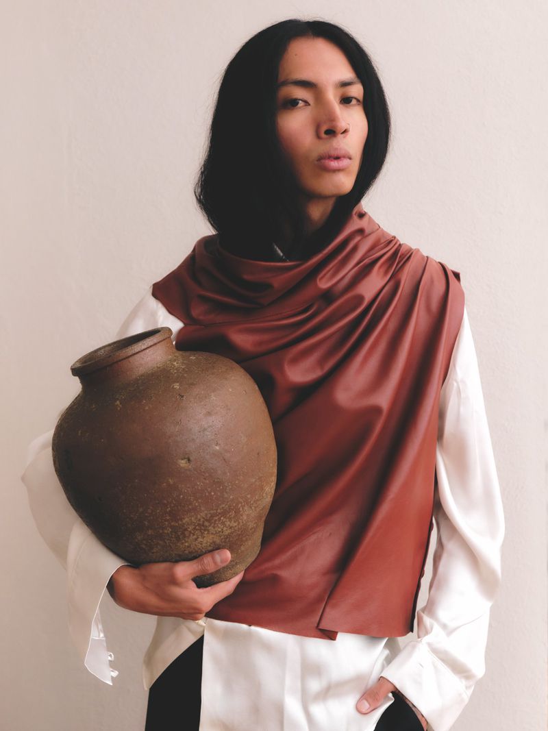 Adult male with long dark hair holding a ceramic vase and draped in a brown cloth stands indoors against a light neutral wall.