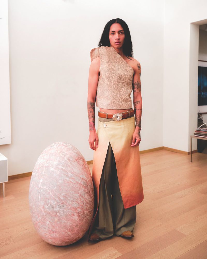 Person in a beige top and layered skirts posing next to a large decorative egg in an indoor setting with neutral colors and modern furniture.