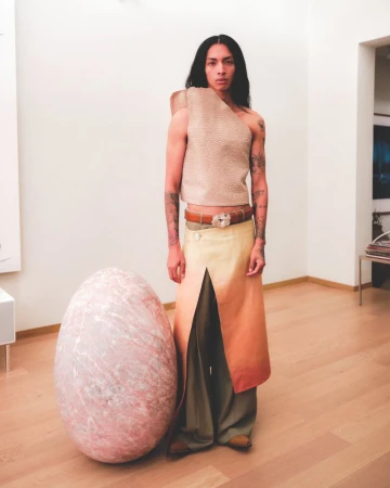 Person in a beige top and layered skirts posing next to a large decorative egg in an indoor setting with neutral colors and modern furniture.