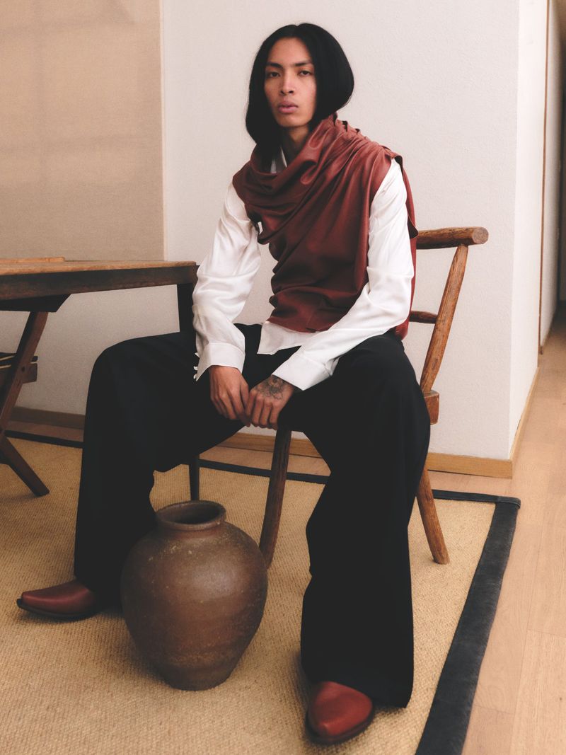 Model wearing a white shirt, red draped top, and black trousers seated on a wooden chair next to a large ceramic pot.