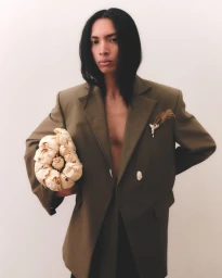 Model with long dark hair wearing an olive blazer and holding a textured beige bundle against a plain background