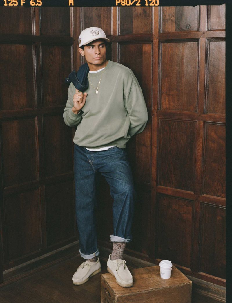 Man in baseball cap and sweatshirt stands indoors against wood paneling with coffee cup on wooden crate, creating casual fashion shot.