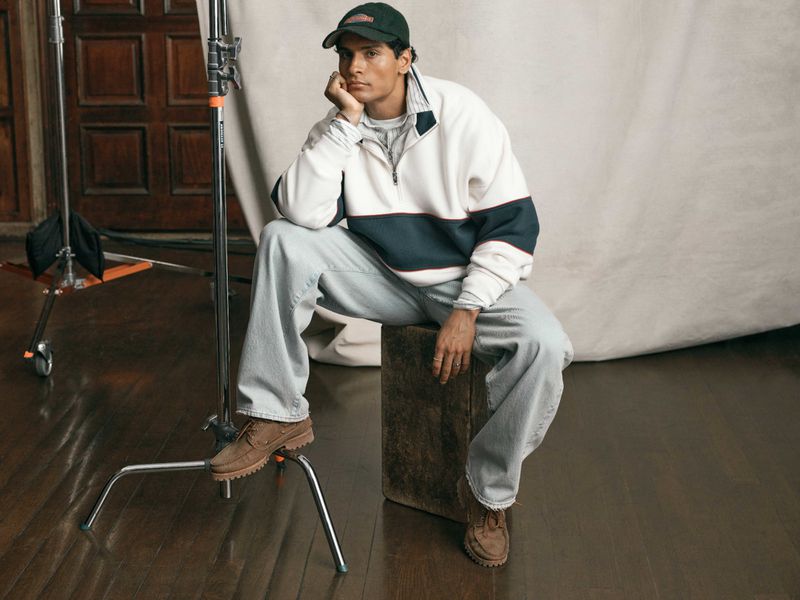 Model seated on wooden crate wearing white quarter-zip sweatshirt, light blue jeans, and dark baseball cap in studio.