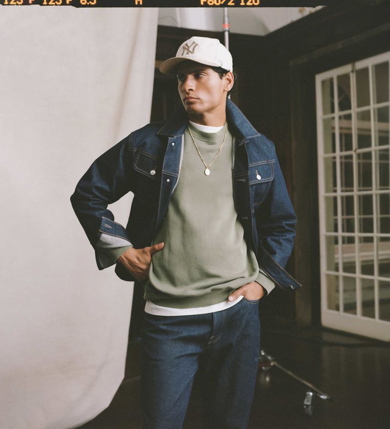 A stylish adult male poses indoors wearing a blue denim jacket, a green sweater, blue jeans, and a baseball cap with a logo.